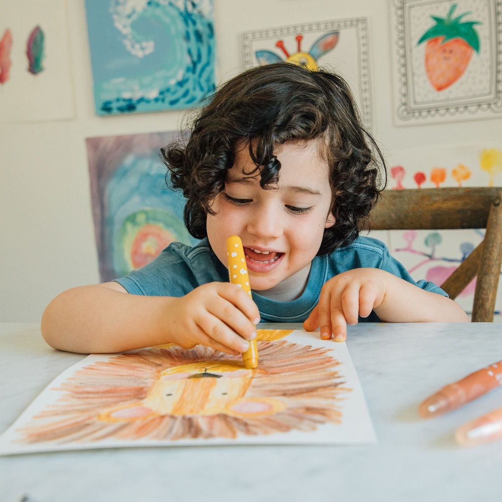 A young child with curly dark hair sits at a table, smiling while coloring a sunburst design on paper with a yellow marker. The scene is set in a brightly lit room with framed artwork on the wall behind them.
